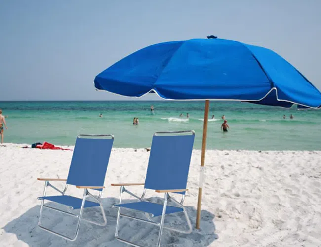 Two blue beach chairs under a matching umbrella on white sand with people swimming in turquoise ocean.