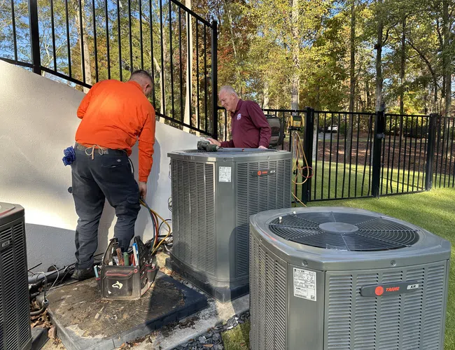 Two HVAC technicians repairing outdoor air conditioning units near a fenced backyard on a sunny day