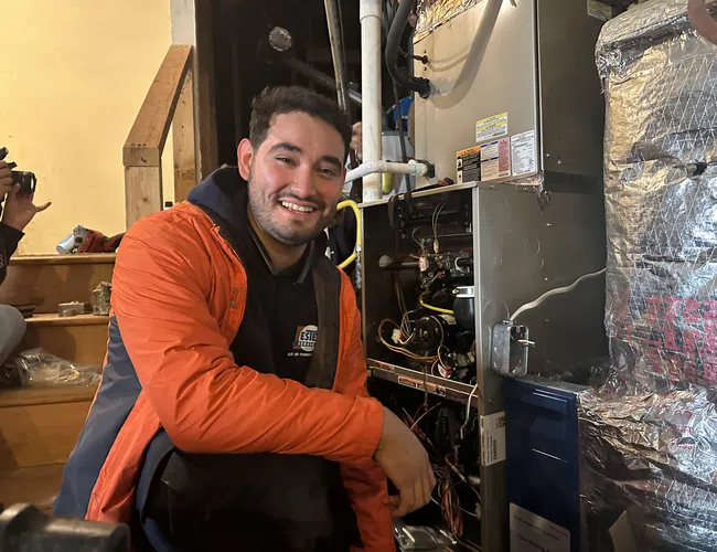 Estes Technician in orange jacket smiling while repairing HVAC system in a residential basement with tools nearby.