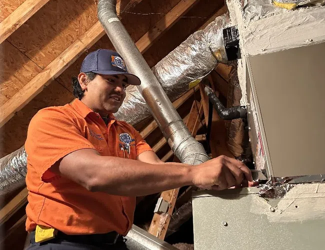 Estes Technician in orange uniform repairing HVAC system ductwork in attic with wooden beams and insulated pipes