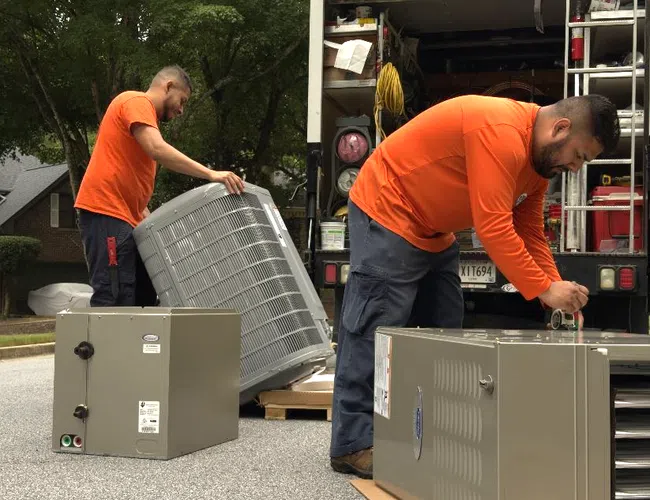 Two Estes technicians in orange shirts loading HVAC units from a truck on a residential street.