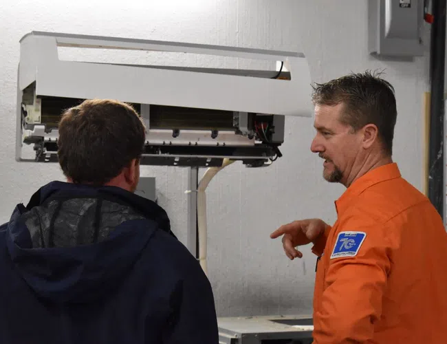 Estes Technician demonstrates air conditioning repair process to a client in a technical workshop.