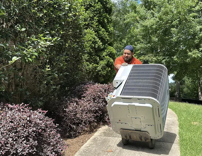 Estes Technician moving a large HVAC unit on a dolly along a garden path surrounded by green trees and shrubs.