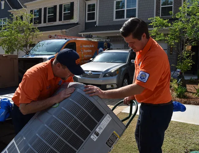 Two Estes technicians installing an air conditioning unit outside a house on a sunny day.