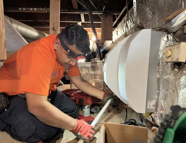 Estes Technician in orange shirt working on HVAC system in a crawl space with tools and equipment around.