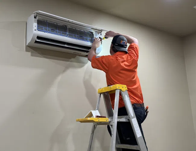 Estes Technician in orange shirt on ladder repairs or installs a wall-mounted air conditioner unit indoors.