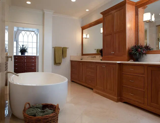 Modern bathroom with freestanding bathtub, wooden cabinets, large mirror, beige tile floor, and green towels.