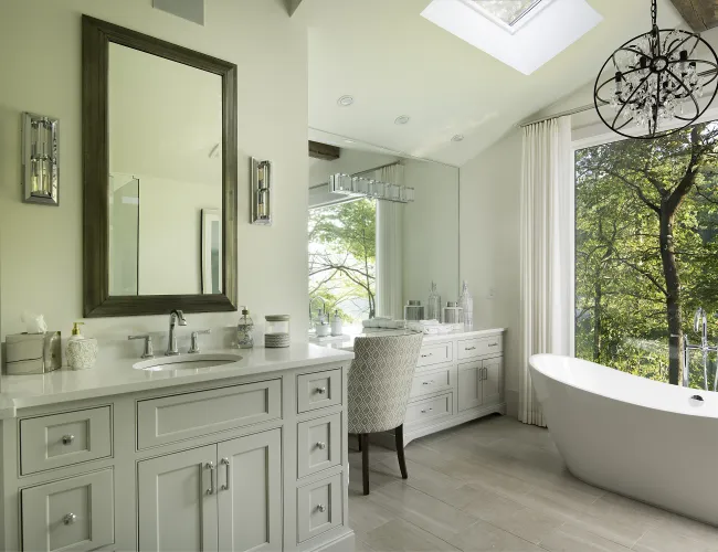 Modern bathroom with white vanity, large mirror, freestanding tub, skylight, and forest view through window.