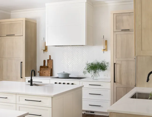 Modern white kitchen with wooden cabinets, marble countertops, dual black faucets, and green plant decoration.