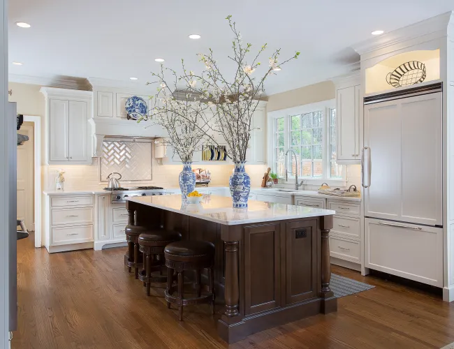 Bright kitchen with white cabinetry, dark wood island, marble countertop, and blue floral vases with branches.
