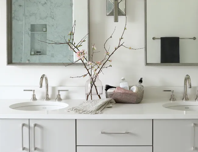 Modern white double vanity with chrome faucets, two mirrors, a floral vase, and countertop toiletries in a minimalist bathroom.