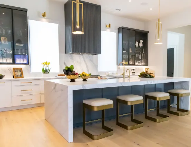 Modern kitchen with large marble island, four bar stools, dark cabinetry, and gold pendant lights over hardwood floor.