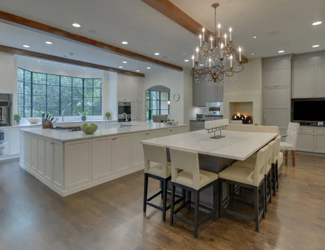 Modern spacious kitchen with large marble island, dining area, chandelier, wood beams, and wide windows overlooking greenery.