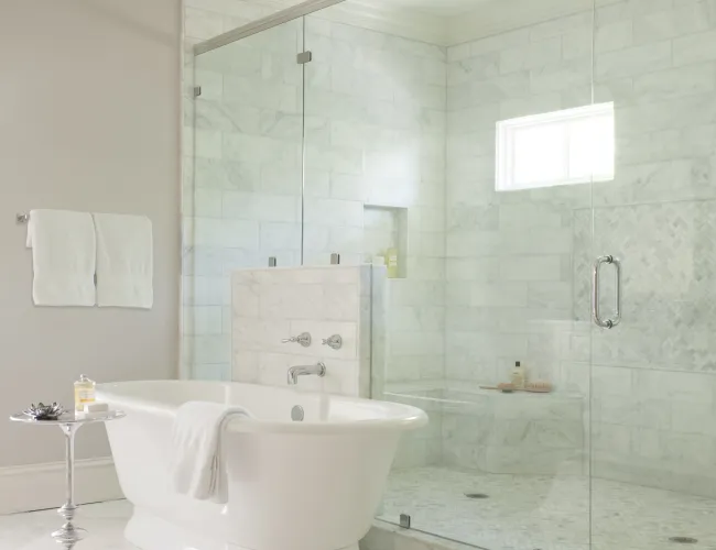 Modern bathroom with white freestanding tub, glass shower enclosure, marble tiles, and soft natural light.