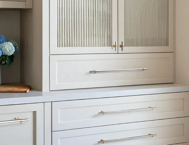 White kitchen cabinetry with fluted glass doors, gold handles, quartz countertop, and a vase of blue hydrangeas.