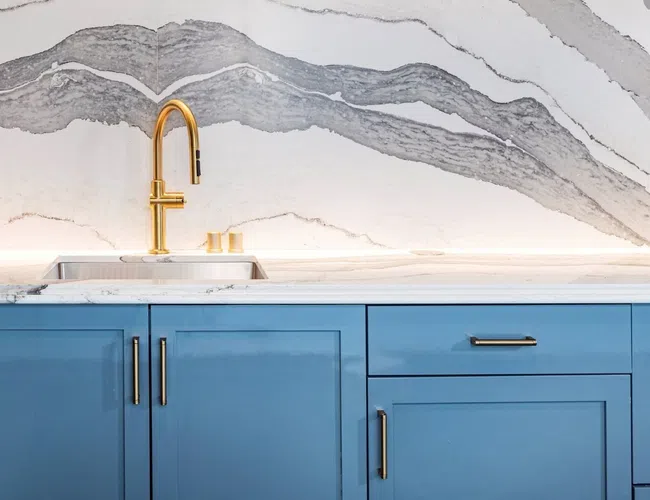 Modern kitchen sink with gold faucet and blue cabinets below white marble countertop and backsplash.
