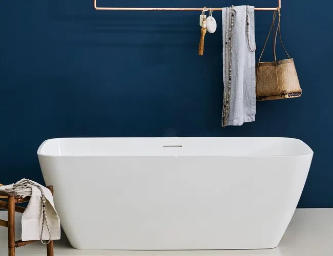 Modern white freestanding bathtub against dark blue wall with hanging towel and basket accessories.