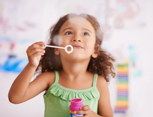Young girl in green top blowing bubbles indoors with colorful artwork in the background, smiling joyfully.