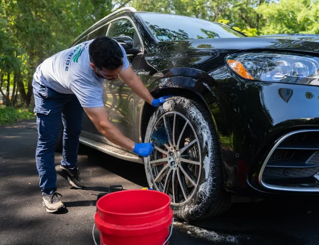 Man wearing gloves washing the tire and rim of a black luxury SUV with soap and water outdoors.