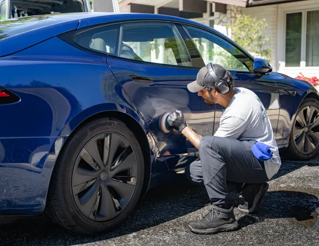 Man polishing the side of a blue electric car outdoors in a residential driveway on a sunny day