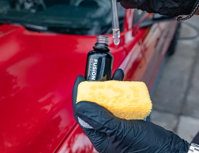 Person wearing black gloves holding ceramic coating bottle and yellow applicator pad near a red car.