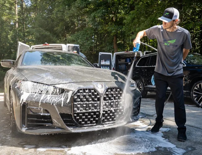 Man washing a black luxury car with soap and water in an outdoor driveway on a sunny day
