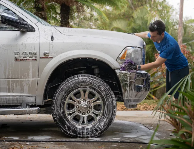 Man washing a white RAM 2500 pickup truck with soap and sponge outdoors surrounded by greenery.