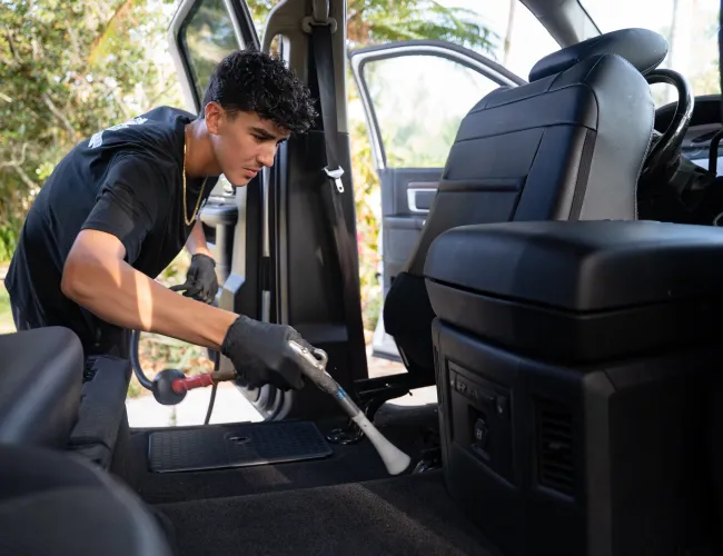 Young man vacuuming the carpet inside a car with open doors on a sunny day outdoors