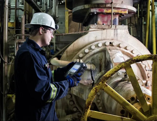 Industrial worker in safety gear using diagnostic device to inspect large machinery in factory setting.