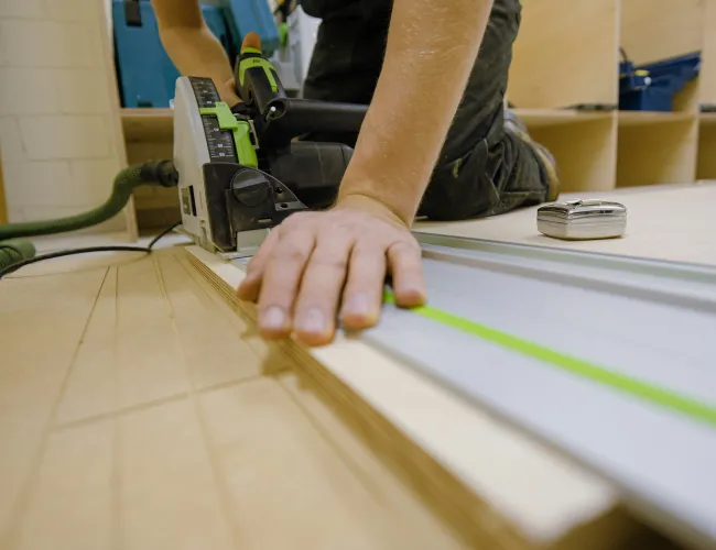 Carpenter using a circular saw with a guide rail to cut plywood on a workshop floor.