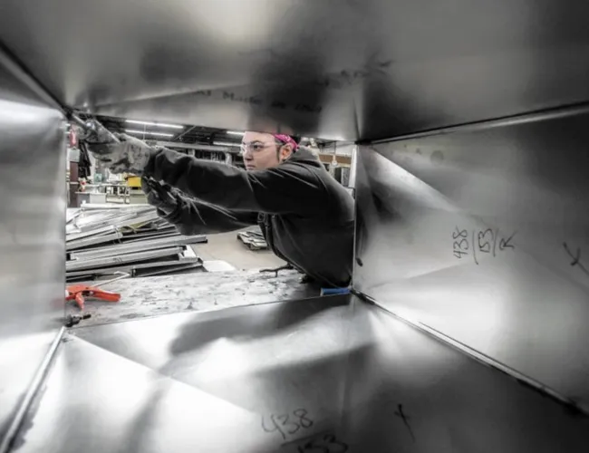 Worker assembling or inspecting metal ductwork inside a manufacturing facility with safety glasses and gloves.