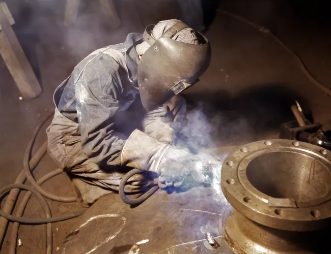 Welder in protective gear welding a large metal pipe in an industrial workshop with sparks and smoke.
