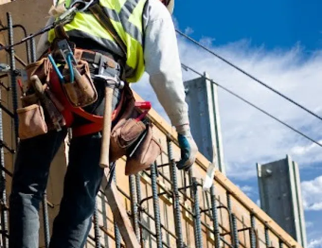 Construction worker in safety gear working on reinforced concrete framework against blue sky.