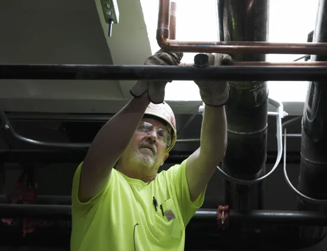 Construction worker wearing safety gear fixing pipes in an industrial setting with overhead lighting.