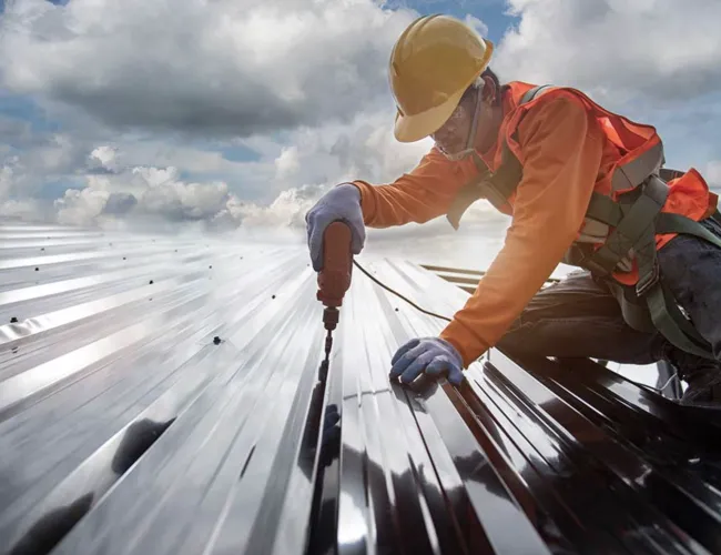 Construction worker in safety gear drilling screws into metal roof panels on a cloudy day.