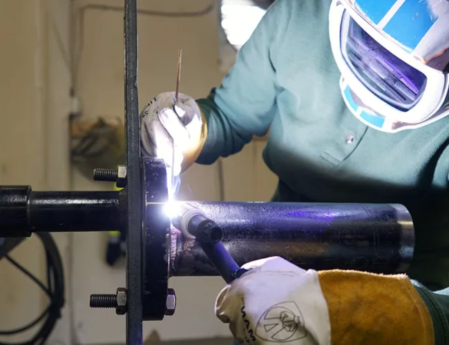 Welder wearing protective gear welding a metal pipe in an industrial workshop setting with bright sparks.