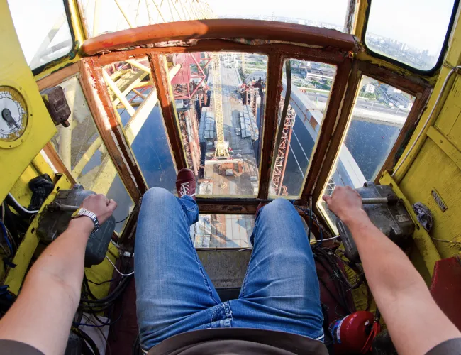 View from inside a crane cabin showing operator's legs and hands controlling machinery high above a construction site.