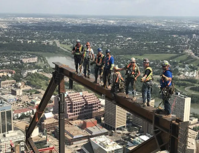 Construction workers in safety gear standing on a steel beam high above a cityscape during building work.