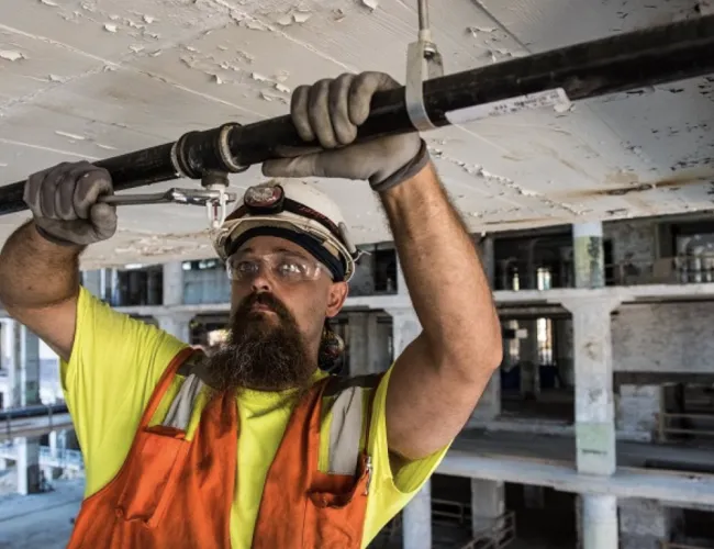 Construction worker in safety gear inspecting pipe in an industrial building under renovation.