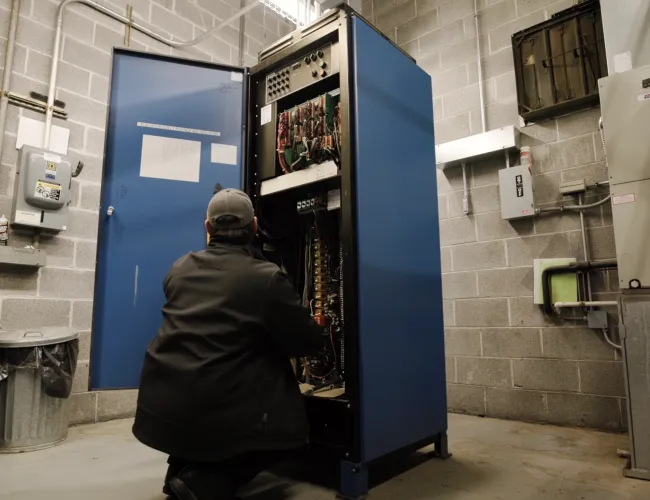 Technician repairing a large blue electrical control panel in an industrial utility room with concrete walls.