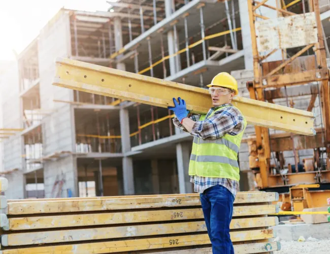 Construction worker carrying yellow metal beam at building site wearing safety gear and helmet