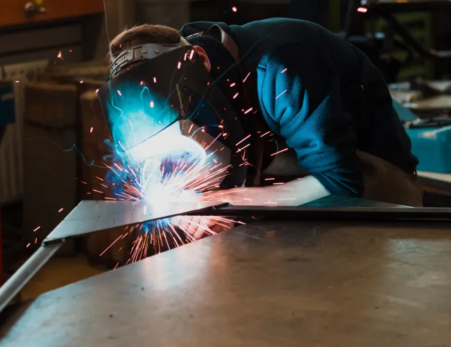 Person welding metal with protective mask, sparks flying in a workshop environment under focused lighting