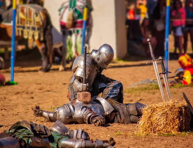 Two knights in armor reenacting a medieval battle scene with swords and hammer on sandy ground outdoors.