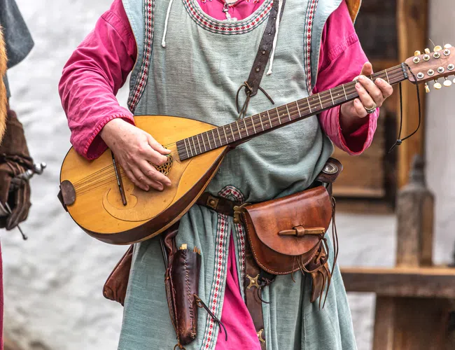 Person in medieval costume playing a stringed lute instrument with leather accessories and a red tunic.