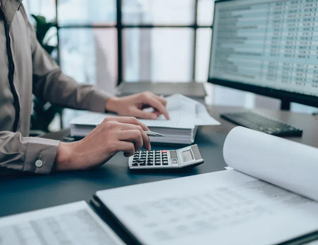 Person calculating finances with a calculator and spreadsheets in a modern office setting.