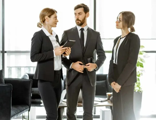 Three professionals in business attire standing and discussing in a modern office with large windows and minimal furniture