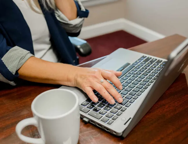 Close-up of hands typing on a laptop keyboard next to a white coffee mug on a wooden desk.