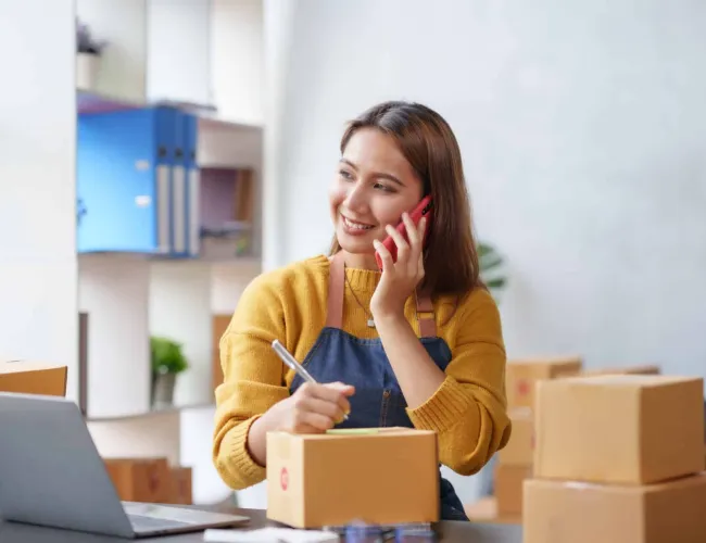 Young woman in yellow sweater and apron talking on phone while packaging boxes in a bright office.