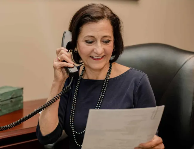 Middle-aged woman sitting at desk talking on phone while reading a document in office setting