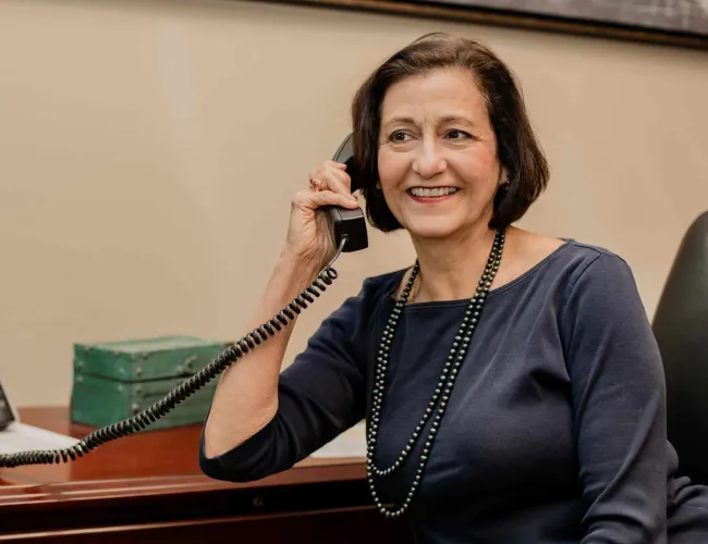 Smiling mature woman talking on a corded office phone at her desk wearing a navy blue top and black beaded necklace